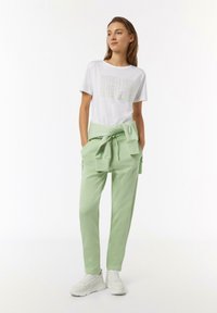 Weißes Baumwoll-T-Shirt mit dem Aufdruck "MINDFULNESS" in wiederholtem Muster; hellgrüne Sweatpants mit gebundenem Bund und seitlichen Taschen. Weiße Turnschuhe.