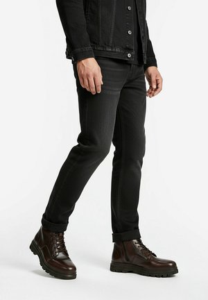 Mann trägt schwarze Jeans mit Umschlägen, schwarze Jeansjacke und braune Leder-Schnürstiefel, steht vor weißem Hintergrund.
