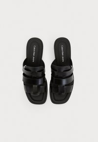 Sandales thématiques noires avec des larges brides, fabriquées dans un matériau lisse. Elles présentent une semelle plate avec des bords texturés et un logo imprimé sur le pied.