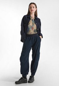 Marineblaues Button-up-Jacket über einem mehrfarbigen Batik-Oberteil, kombiniert mit dunklen Cargo-Hosen und schwarzen Sneakers. Lässiger Schnitt; weiche Texturen.