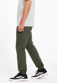 Pantalones de chándal verde olivo con puños elásticos, bolsillos laterales y un acabado mate suave, combinados con zapatillas negras y blancas.