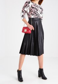 Schwarzer plissierter Midi-Rock kombiniert mit einem weißen Oberteil mit pinken Blumenmustern. Eine kleine rote Handtasche und schwarze Stiefeletten runden das Outfit ab.
