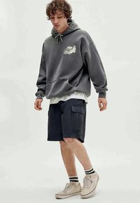 Grijze hoodie met een voorzak en grafisch ontwerp, gecombineerd met donkere cargoshorts. Lichtgekleurde sneakers en gestreepte sokken maken de outfit compleet.