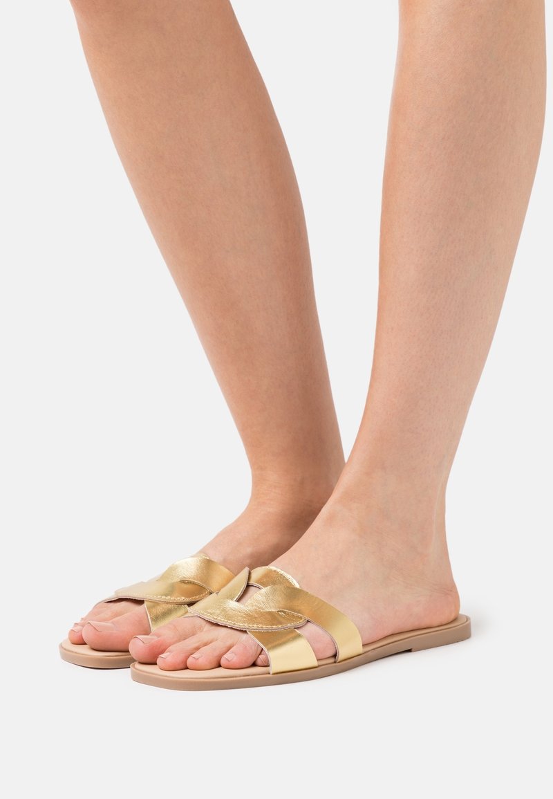 Sandales glissantes dorées avec un design de sangle torsadée, semelle plate et texture lisse. La partie supérieure présente des accents métalliques brillants et une base neutre.