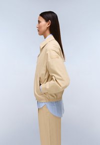 Kobieta w beżowej kurtce i spodniach z niebieską koszulą w paski pod spodem stoi bokiem na tle jasnego tła.