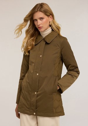 Giacca trapuntata verde oliva con colletto arrotondato, bottoni a pressione, tasche laterali e una texture liscia. Indossata sopra un dolcevita color crema.