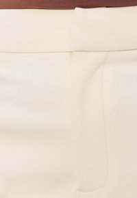 Gros plan sur un pantalon de couleur crème avec des coutures détaillées et une ceinture, partiellement couvert par un haut marron.