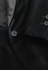 Blazer in velluto scuro con una morbida fodera interna, due bottoni e dettagli di cucitura a contrasto. Esterno strutturato con una leggera lucentezza.