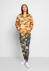 Kamuflasjefarget jakke i oransje, gule og grønne toner; glidelås foran, hette. Kamuflasje leggings i grønne, brune og blå nyanser. Hvite sneakers.