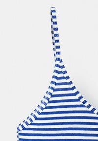 Gros plan d'une bretelle de maillot de bain en tissu rayé bleu et blanc horizontalement et de la partie supérieure de la poitrine sur un fond uni.