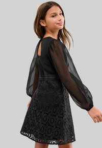 Robe noire avec un blouse transparente à manches longues et une taille smockée, ornée de détails en dentelle et de motifs circulaires sur la jupe.