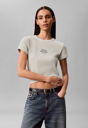 Kvinde med langt mørkt hår iført en lys grå Calvin Klein crop t-shirt og mørkeblå jeans med sort bælte, stående mod en ensfarvet grå baggrund.