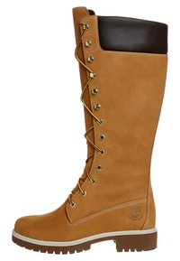 Bota impermeable de caña alta en color marrón claro con puño de cuero marrón, herrajes dorados y diseño de cordones. Acabado en suede texturizado con suela de goma.