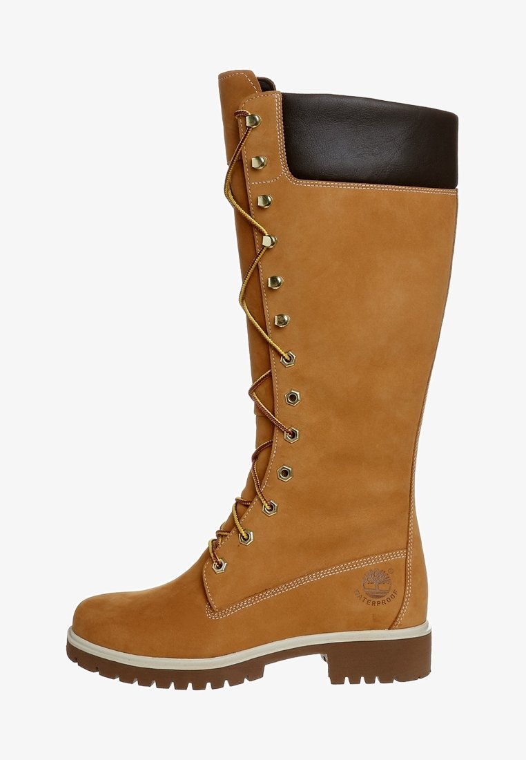Bota impermeable de caña alta en color marrón claro con puño de cuero marrón, herrajes dorados y diseño de cordones. Acabado en suede texturizado con suela de goma.