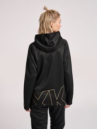 Vrouw met blond haar in een korte paardenstaart, die een zwarte hoodie en broek draagt met gele geometrische lijnpatronen op de achterkant.