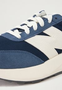 Plave čizme od suede i mrežastog materijala s teksturiranim bijelim logotipom, ravnim vezicama i jastučićem u đonu. Pogled izbliza koji ističe dizajnerske elemente.