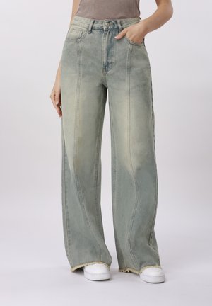 Kobieta ubrana w spodnie z wysokim stanem, szerokie nogawki, wyblakłe zielone z postrzępionymi brzegami, w połączeniu z białymi trampkami, stojąca z jedną ręką w kieszeni.