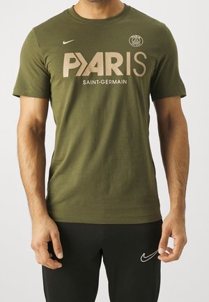 Man draagt een olijfgroen Paris Saint-Germain T-shirt en zwarte Nike broek, staat met ontspannen armen langs zijn zij.