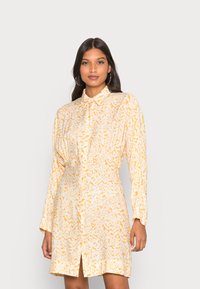 Vestido de manga longa amarelo e branco com colarinho abotoado, apresentando um padrão de leopardo texturizado e uma fenda frontal para maior mobilidade.