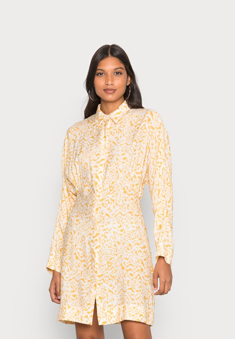 Vestido de manga longa amarelo e branco com colarinho abotoado, apresentando um padrão de leopardo texturizado e uma fenda frontal para maior mobilidade.