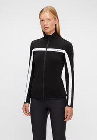Svart zip-jacka med hög krage, prydd med vita ränder på ärmarna och en bröstlogotyp. Tillverkad av ett mjukt, stretchigt tyg.
