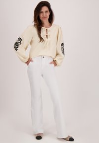 Crème blouse met een strikje aan de voorkant en zwarte bloemborduursels op de mouwen, gecombineerd met witte flairjeans en zwarte flats met een punt.