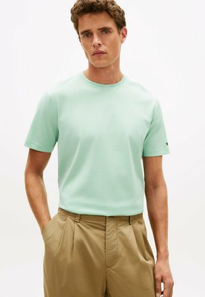 Junger Mann trägt ein hellgrünes kurzärmeliges Hemd, das in eine hoch taillierte khakifarbene Hose mit Falten gesteckt ist, die Hände in den Taschen.