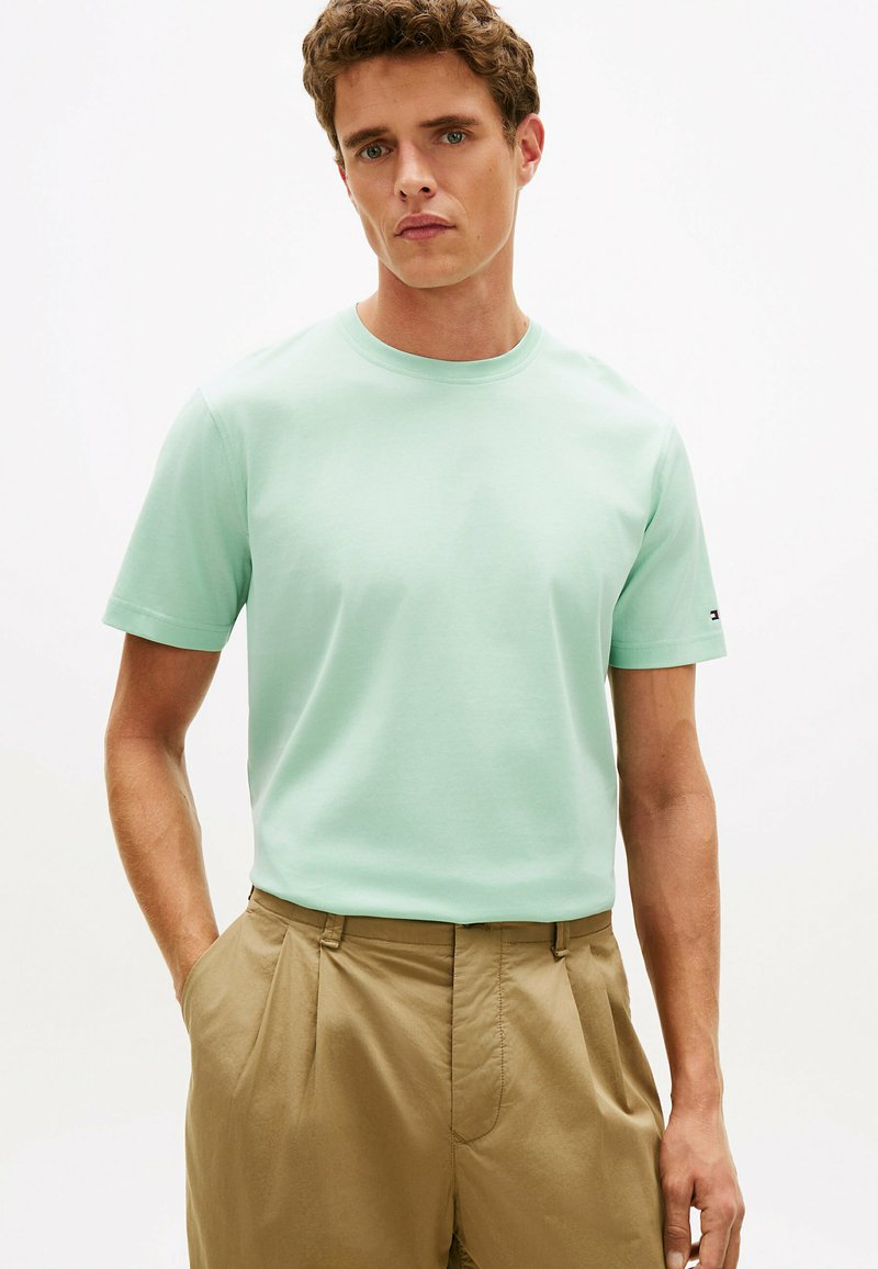 Jeune homme portant une chemise à manches courtes vert clair rentrée dans un pantalon kaki plissé taille haute, les mains dans les poches.