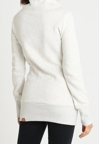 Vaaleanharmaa villapaita, jossa on pitkät hihat, korkea kaulus, ribbaushelma ja teksturoitu kangas. Takana on pieni brändilogo.