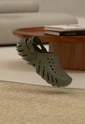 Grøn slip-on træsko med ventilationshuller og justerbar hælrem svæver over beige tæppe i en stue med træbord og bøger.