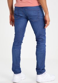 Blå denimjeans med slim fit, med bakfickor och kontrasterande sömmar, i kombination med vita sneakers på en neutral bakgrund.