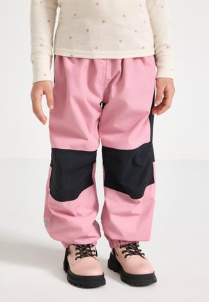 Barn iført lyserøde og sorte vandtætte bukser med elastiske manchetter og lyserøde snørebåndsstøvler på hvid baggrund.