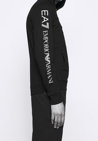 Černá mikina na zip s přední kapsou, s bílým textem "EA7" a "Emporio Armani" na rukávu. Hladká textura a přiléhavý design.