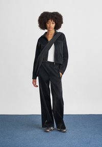 Μαύρο φόρμα από λείο, γυαλιστερό υλικό, με μπουφάν που κλείνει με φερμουάρ και παντελόνι με ελαστική μέση. Συμπληρώνεται με μια υφασμάτινη τσάντα χιαστί.