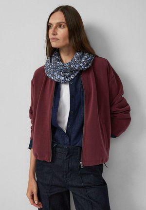 Frau mit langen braunen Haaren, die einen gemusterten blauen Schal, eine kastanienbraune Bomberjacke, ein Jeanshemd und dunkle Jeans trägt, steht vor einer grauen Wand.