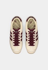 Sneaker crema e bordeaux realizzate in mesh e suede, con tre strisce decorative, punta rotonda e suola in gomma texturizzata.