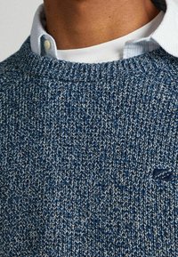 Modro-bílé pletené svetr s texturovaným vzorem, vybavený ribovaným výstřihem a jemným logem na hrudi.