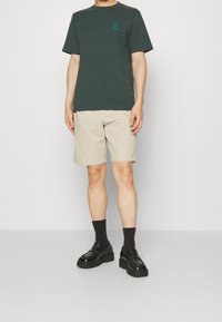 Tmavě zelené bavlněné tričko s malým tyrkysovým logem, spárované se béžovými šortkami, černými ponožkami a robustními černými botami. Model stojí vzpřímeně.