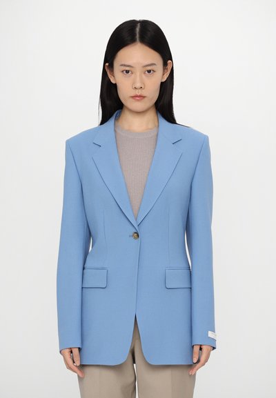 Blazer su misura azzurro chiaro con rever affilati, chiusura a un bottone e due tasche frontali; indossato sopra un top grigio a coste e pantaloni beige.