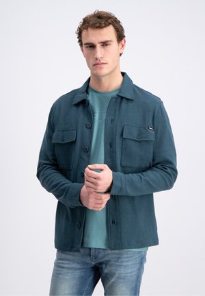 Joven con chaqueta abotonada color verde azulado sobre una camiseta azul verdosa y vaqueros desgastados, de pie frente a un fondo blanco liso.