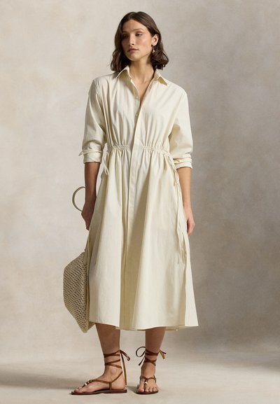 Φόρεμα πουκαμίσα σε ανοιχτό μπεζ χρώμα με κουμπιά, σούρα στη μέση, μακριά μανίκια και πλαϊνές τσέπες. Φορεμένο με καφέ σανδάλια με λουράκια και πλεκτή τσάντα.