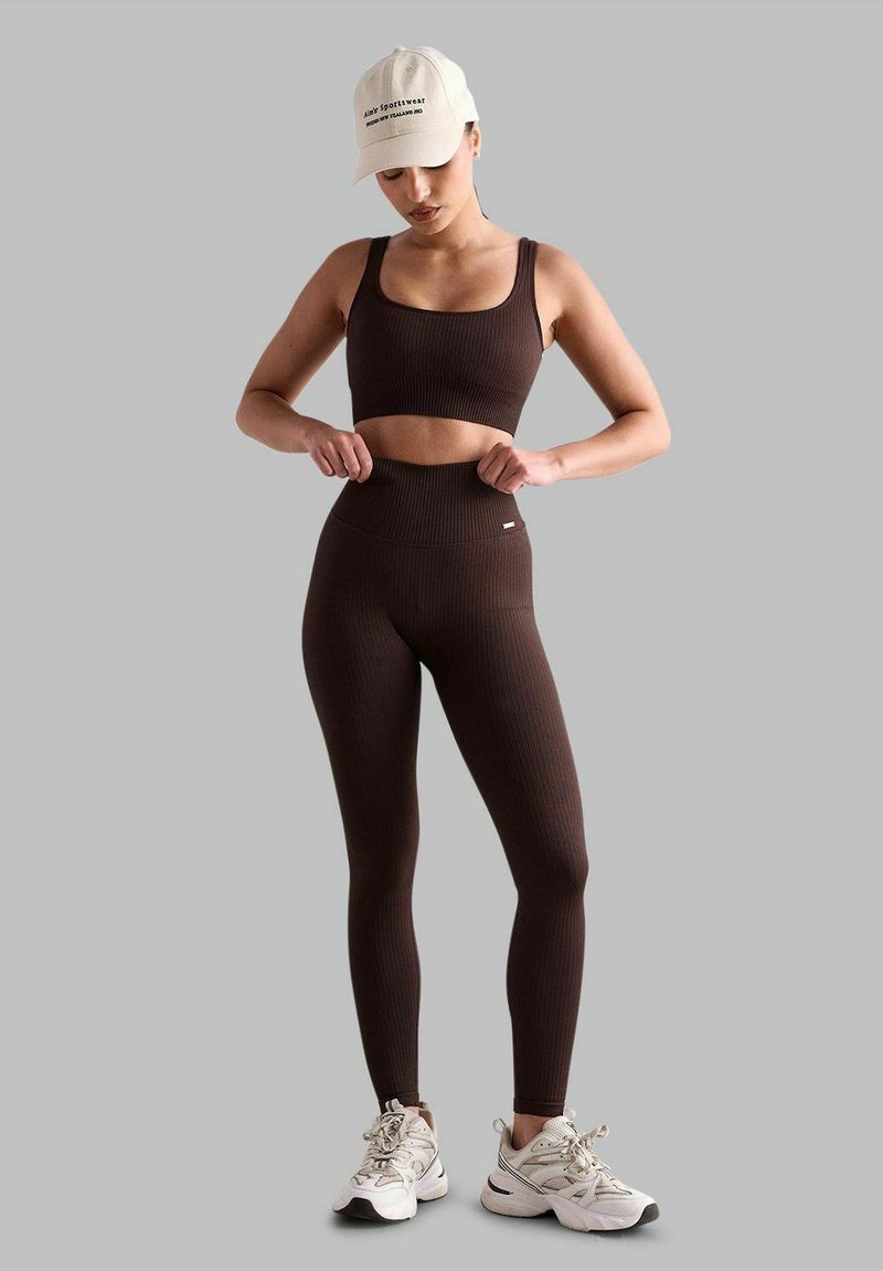 Pruun ribadega spordibrasi ja leggingide komplekt, millel on kõrge vöökoht ja tagasihoidlik logo. Kombineeritud heledabež jacket ja valgete tossudega.