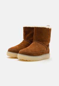 Stivaletti in suede marrone con una morbida fodera in pelliccia color crema, dotati di una suola piatta in gomma traslucida e dettagli di cucitura minimalista.