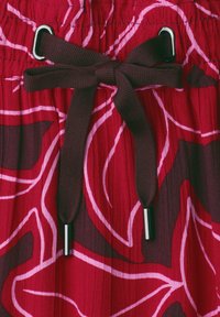 Lacet marron noué en nœud sur un tissu rouge avec des motifs de feuilles roses et rouge foncé et des extrémités argentées.