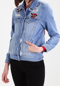 Světle modrá džínsová bunda s poškozenými detaily, dvěma náprsními kapsami, vyšitými nášivkami a slim fit střihem. Zapínání na knoflíky a ohrnuté rukávy.