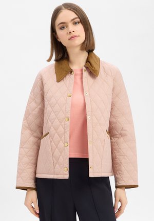 Junge Frau mit glatten braunen Haaren trägt eine hellrosa gesteppt Jacke mit braunem Kordkragen über einem hellrosa Oberteil und schwarzen Hosen.