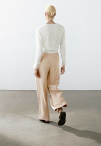 Lichtgewicht, crèmekleurige gebreide trui gecombineerd met een beige hoge taille, brede broek met een subtiel verticaal streepjespatroon. Zwarte platte schoenen.