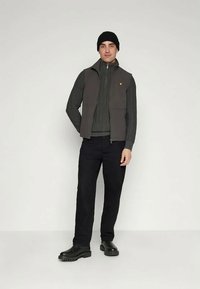 Gilet grigio senza maniche sopra un maglione scuro a coste, abbinato a pantaloni neri e stivali neri. Un piccolo logo giallo è visibile sul gilet.