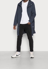 Man med en marinblå huva över en vit grafisk t-shirt, svarta jeans, vita strumpor och vita Nike-sneakers med svart swoosh.