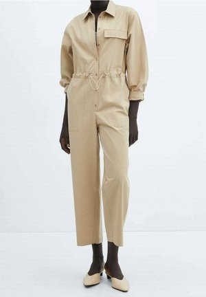 Model wearing beige long-sleeve jumpsuit with drawstring waist, chest pocket, and beige low-heeled shoes against white background.